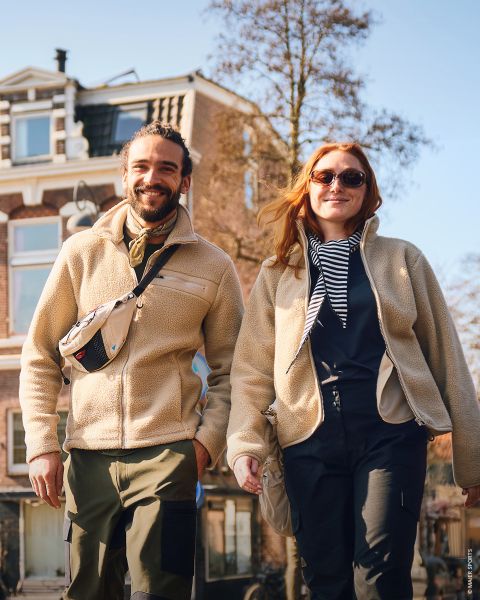 Un homme et une femme souriants se promènent en plein air par une journée ensoleillée, vêtus de vestes et de bottes décontractées, se tenant la main, avec un grand bâtiment en briques en arrière-plan.