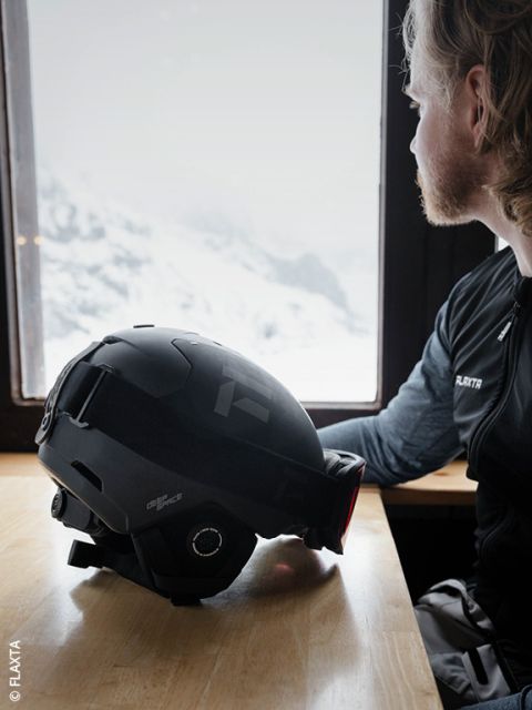 Une personne aux cheveux clairs et à la barbe est assise à une table en bois près de la fenêtre et regarde les montagnes enneigées à l'extérieur. Un casque de ski noir est posé devant elle sur la table.