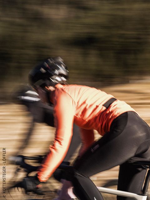 Un cycliste portant une veste Löffler verte et un casque regarde droit devant lui avec un air concentré. L'image est légèrement floue, ce qui met en valeur les détails du haut du corps et de la veste.