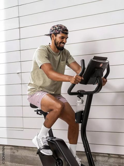 Un homme portant une casquette à fleurs, un t-shirt beige et un short rose sourit en faisant du vélo d'appartement Schwinn.