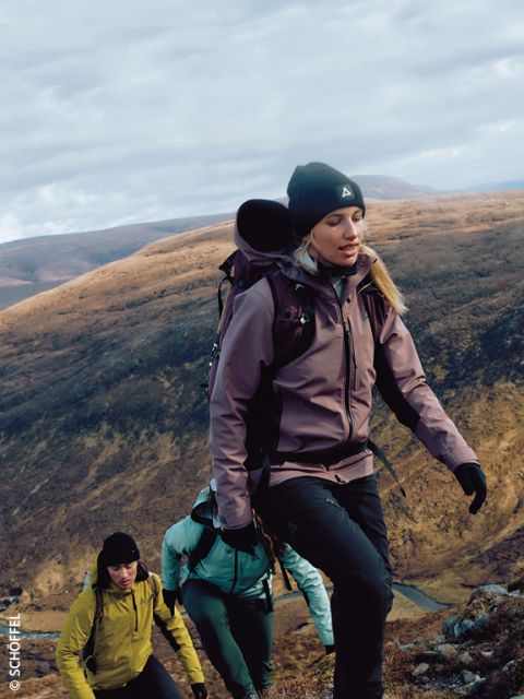 Trois randonneurs équipés de matériel de plein air et de sacs à dos gravissent une pente rocheuse avec des collines douces et brunes en arrière-plan sous un ciel nuageux. Le randonneur en tête porte une veste violette et un bonnet.