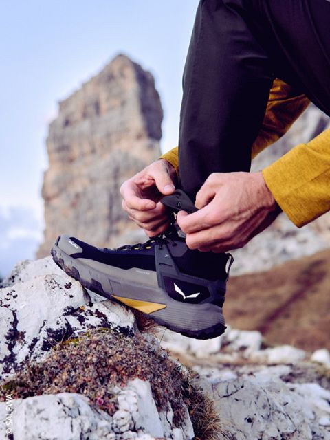 Une personne en tenue de plein air serre les lacets d'une chaussure de randonnée noire, debout sur une surface rocheuse, avec une grande formation rocheuse et un ciel bleu en arrière-plan.
