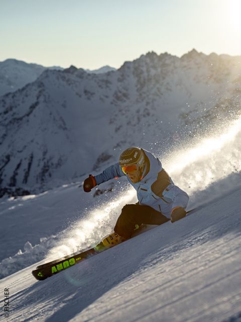 Un skieur en veste bleue et avec un casque dévale une pente de montagne enneigée au coucher du soleil, soulevant de la poudreuse, avec des sommets montagneux escarpés et ensoleillés en arrière-plan.
