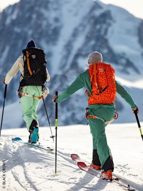 Deux personnes en vêtements de ski colorés et avec des sacs à dos montent une montagne enneigée en s'aidant de bâtons. En arrière-plan, on aperçoit un grand sommet montagneux escarpé.
