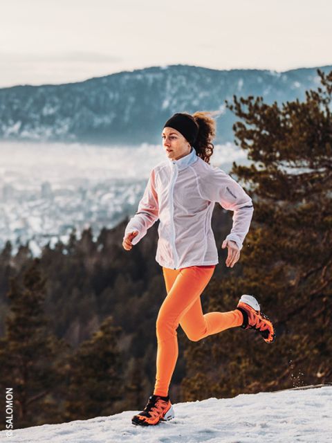 Une femme vêtue d'une veste blanche et de leggings orange vif court sur un chemin enneigé avec des pins et des montagnes en arrière-plan. Le ciel est nuageux et le paysage en contrebas est partiellement recouvert de neige.