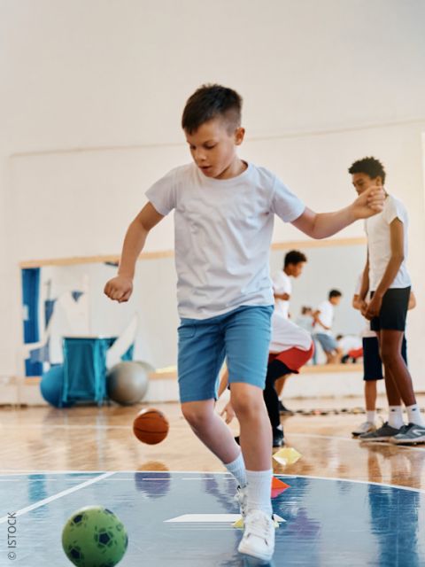 Un garçon en t-shirt blanc et short bleu est en équilibre sur un pied pendant qu'il joue dans un gymnase. On aperçoit d'autres enfants et des ballons de sport en arrière-plan.