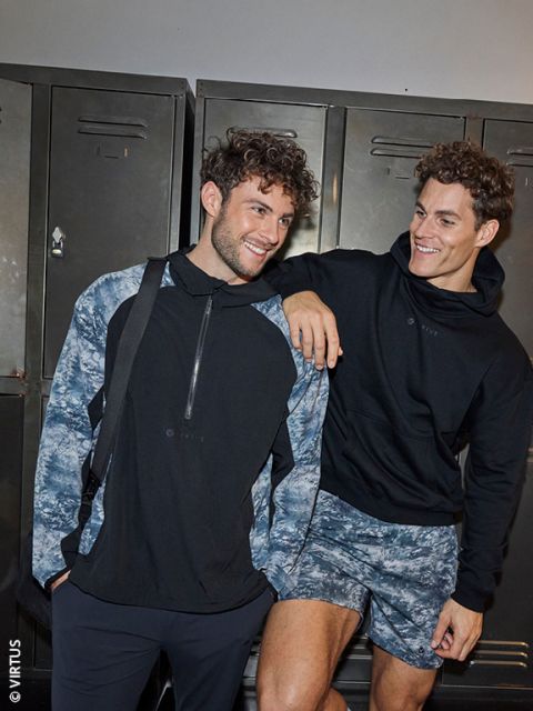 Deux jeunes hommes en tenue de sport sourient et posent ensemble devant un casier. L'un porte une veste noire et bleue et un sac à bandoulière, l'autre un sweat à capuche noir et un short à motifs et pose son bras sur l'épaule de son ami.