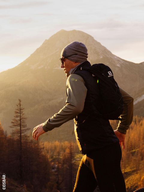 Une femme portant une veste et des gants rose-gris court dans un paysage montagneux enneigé, transportant deux bouteilles d'eau dans les poches avant de son gilet de course.
