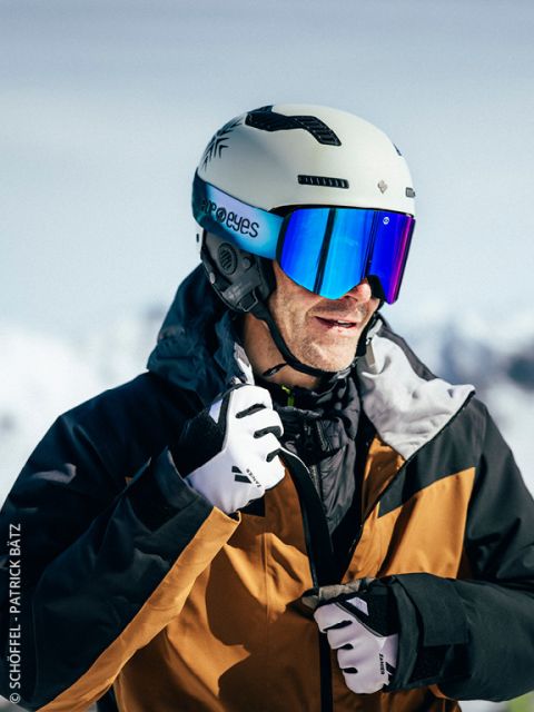 Un skieur portant un casque blanc, des lunettes réfléchissantes, une veste et des gants noir et marron ferme sa veste à l'extérieur avec des montagnes enneigées en arrière-plan.