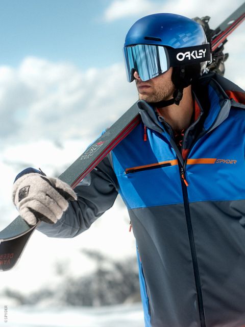 Un skieur en veste bleue, casque et lunettes de ski porte des skis sur l'épaule devant un paysage de montagne enneigé.
