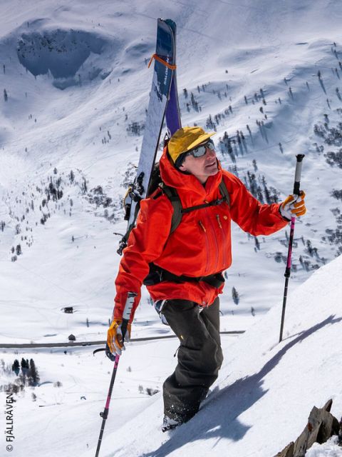 Une personne en veste rouge et bonnet jaune escalade une pente de montagne enneigée, porte des skis sur son sac à dos et tient des bâtons de ski à la main.