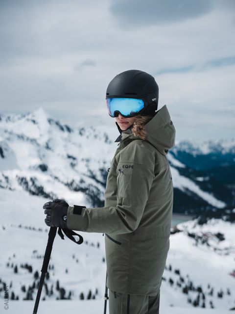Un skieur en veste verte et casque noir se tient sur une pente de montagne enneigée, tenant des bâtons de ski et regardant en arrière, avec des sommets en arrière-plan.