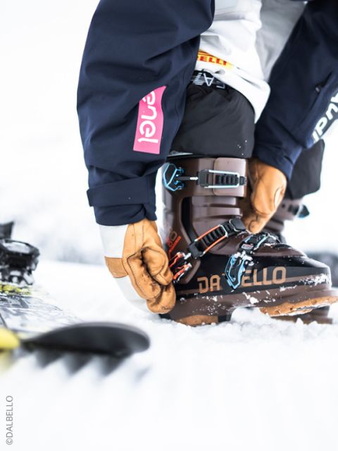 Un skieur portant des gants ferme les boucles d'une chaussure de ski Dalbello noire, avec des skis et de la neige en arrière-plan.