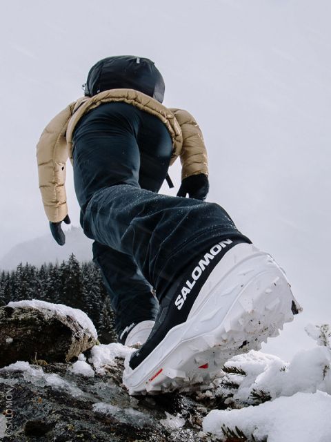 Une personne en vêtements d'hiver et chaussures Salomon monte une pente enneigée, l'accent étant mis sur la semelle de la chaussure, avec une montagne enneigée en arrière-plan.