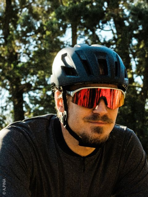 Un homme avec un casque de vélo noir, des lunettes de soleil rouges réfléchissantes et une chemise foncée se tient à l'extérieur avec des arbres en arrière-plan.