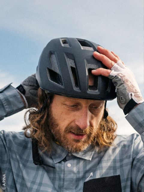 Un homme aux cheveux longs et à la barbe ajuste son casque de vélo noir, il porte une chemise à carreaux et des gants sans doigts, le ciel est visible en arrière-plan.