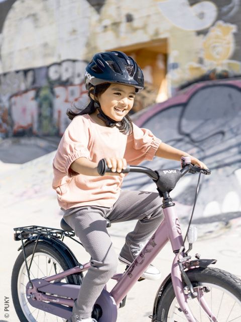 Une jeune fille souriante avec un casque fait du vélo rose devant un mur couvert de graffitis.