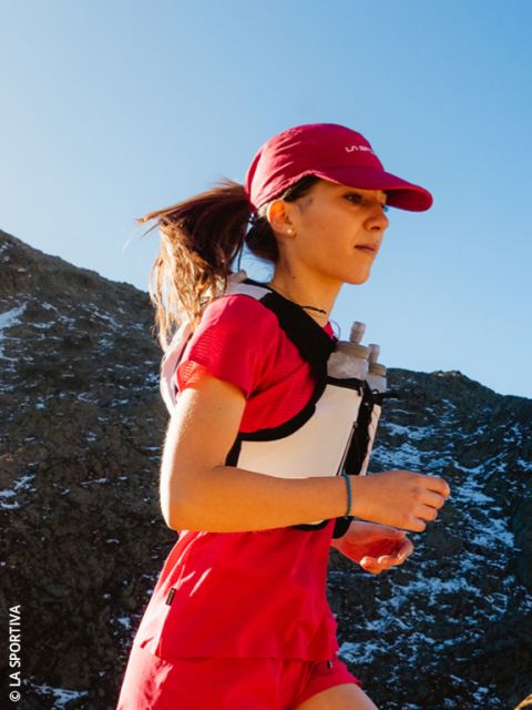 Une femme en tenue de course rouge fait du jogging avec un gilet d'hydratation sous un ciel bleu sur un parcours de montagne.