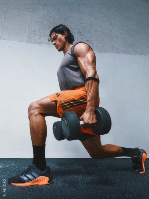 Un homme musclé en tenue de sport effectue une fente avec des haltères dans une salle de sport et porte des baskets Adidas noires et orange.
