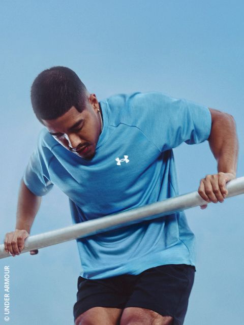 Un homme portant un t-shirt bleu Under Armour effectue un exercice de dips sur des barres parallèles devant un ciel bleu éclatant.