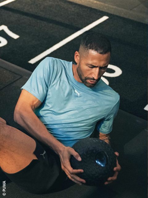 Un homme portant un t-shirt Puma bleu clair s'entraîne sur le sol d'une salle de sport et tient un ballon de médecine noir texturé à deux mains.