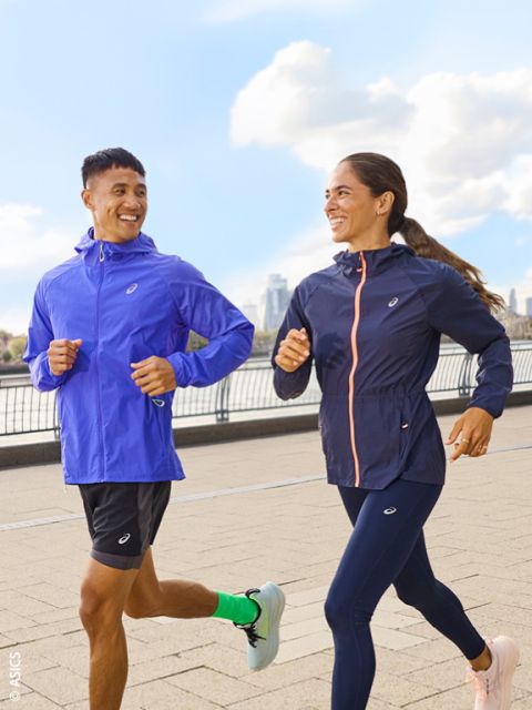 Deux personnes en tenue de sport font du jogging à l'extérieur sur un chemin pavé au bord d'une rivière et se sourient, avec la ligne d'horizon de la ville et le ciel bleu en arrière-plan.