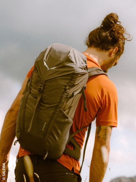 Une personne avec un chignon, une chemise orange et un sac à dos se tient à l'extérieur sous un ciel nuageux et regarde au loin.