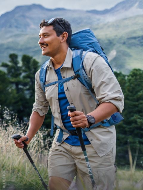 Un homme, faisant de la randonnée dans la nature avec des bâtons de marche, portant un sac à dos, une chemise beige et un short, et souriant sur fond d'arbres verts et de montagnes.