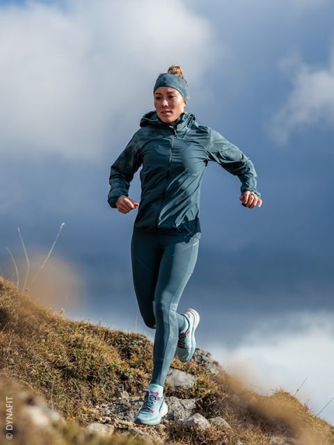 Une femme en tenue de sport turquoise court en descente sur un sentier rocheux, 