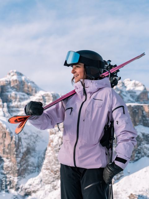 Une skieuse en veste lavande et casque sourit en portant ses skis sur l