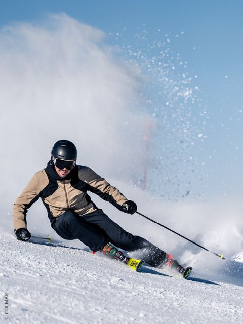 Un skieur avec un casque et une veste marron clair effectue des virages serrés s