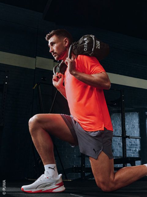 Un homme en T-shirt orange et short gris fait une fente dans une salle de sport,