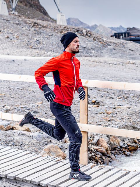 Un homme en veste rouge, pantalon noir, gants et bonnet marche sur un pont en bo